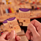 Close-up of hands holding a wooden card with purple top and brick pattern, blurred board game in the background